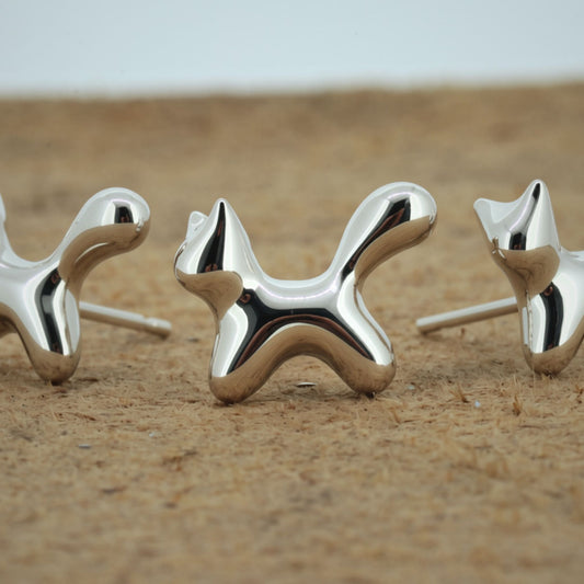 Three polished silver dog-shaped stud earrings displayed on a sandy surface with a blurred light gray background.