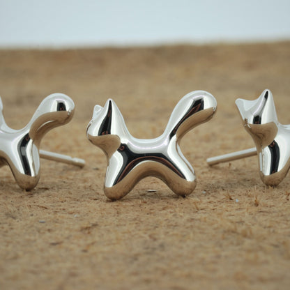 Three polished silver dog-shaped stud earrings displayed on a sandy surface with a blurred light gray background.